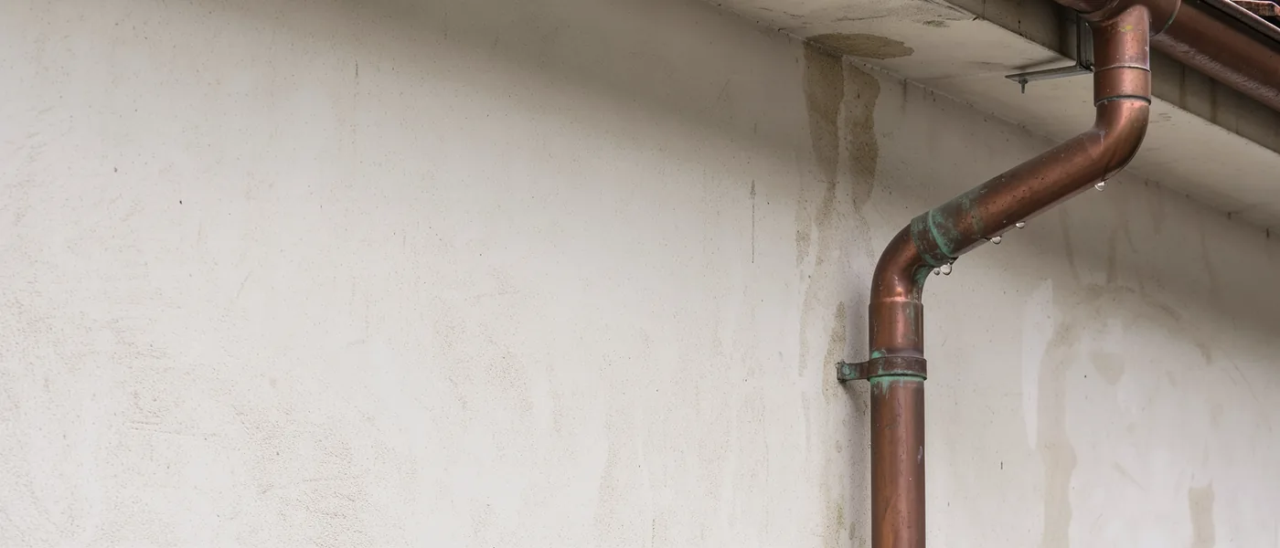 A copper downpipe bends below a roof edge on a pale apartment exterior.